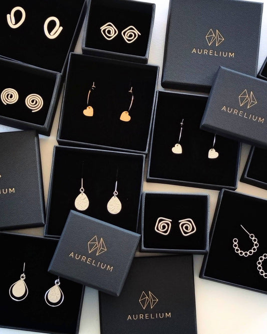 Collection of gold and silver earrings in black square gift boxes with gold geometric logo and the word "Aurelium" in gold lettering on box lids