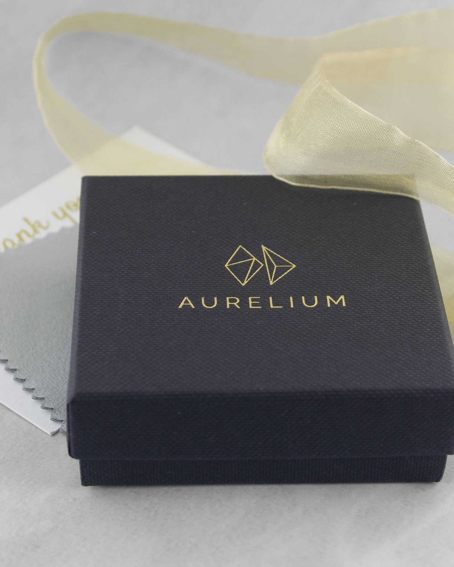 Closed black jewellery gift box with gold logo and the word "Aurelium" printed in gold lettering lying beside gold ribbon and a polishing cloth