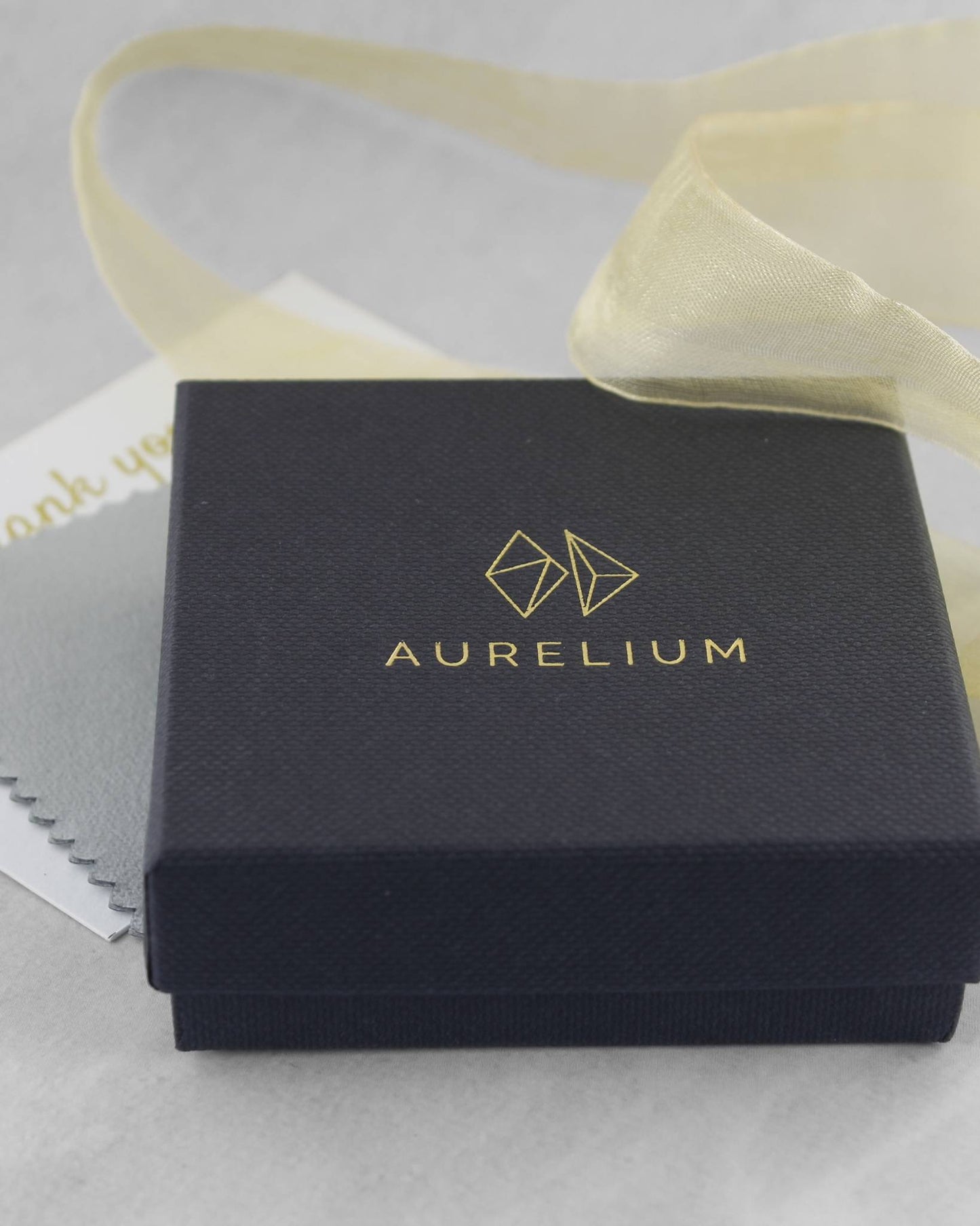 Closed black jewellery gift box with gold logo and the word "Aurelium" printed in gold lettering lying beside gold ribbon and a polishing cloth