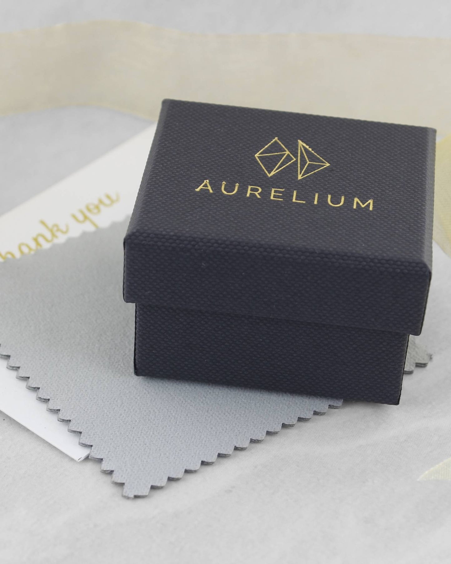 Closed black ring box with gold logo and the word "Aurelium" in gold lettering printed on lid, lying beside polishing cloth and thank you card
