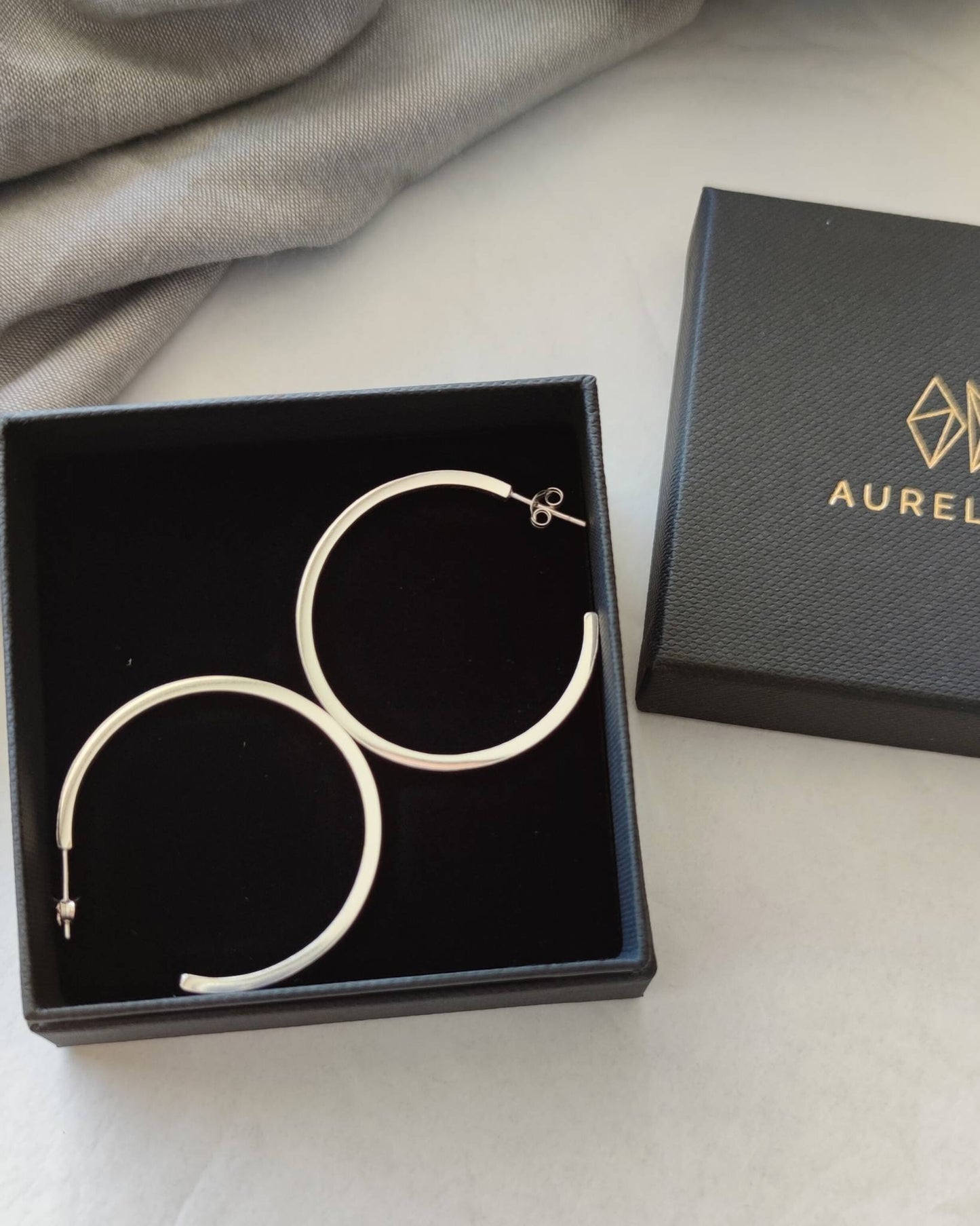 Large sterling silver hoop earrings lying in black jewellery gift box beside box lid printed with Aurelium logo in gold