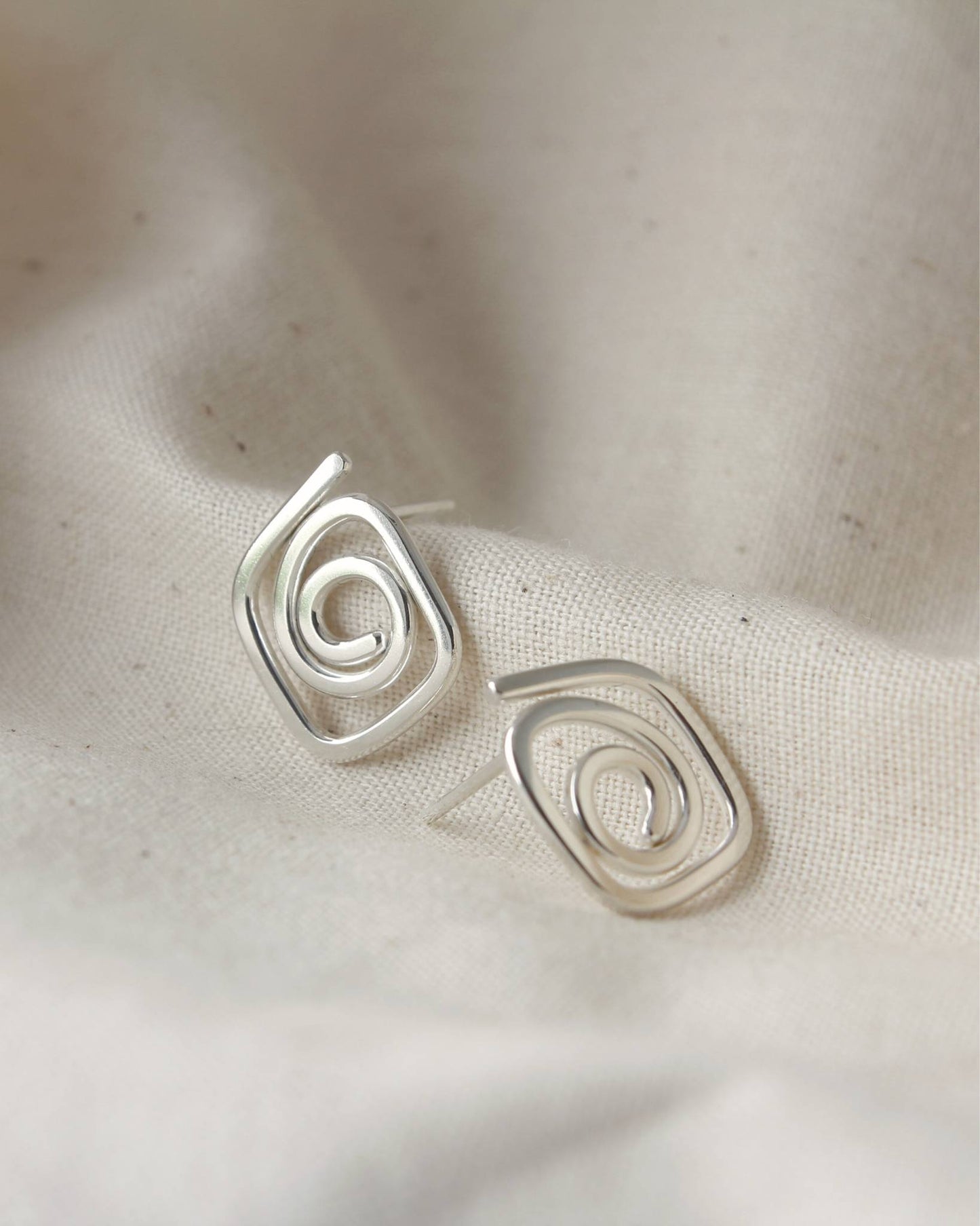 Sterling silver diamond-shaped stud earrings in a swirl design lying on beige fabric