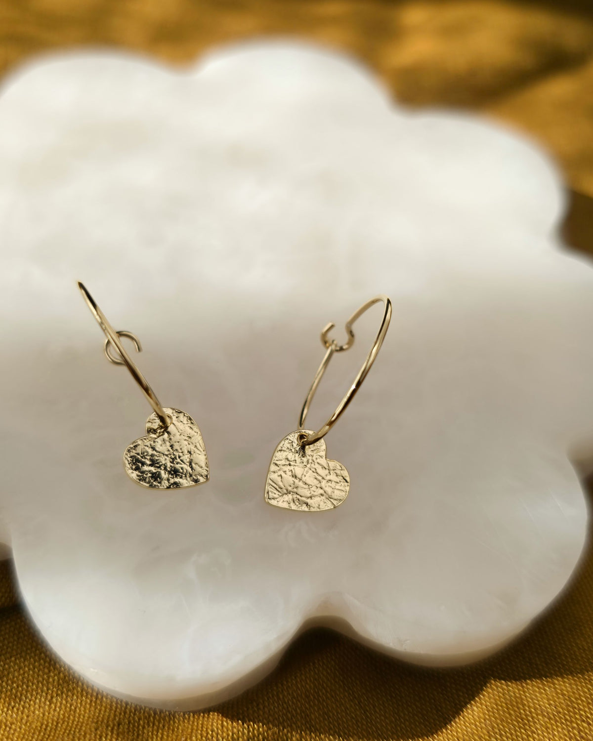 Delicate textured gold heart earrings on cream petal-shaped surface