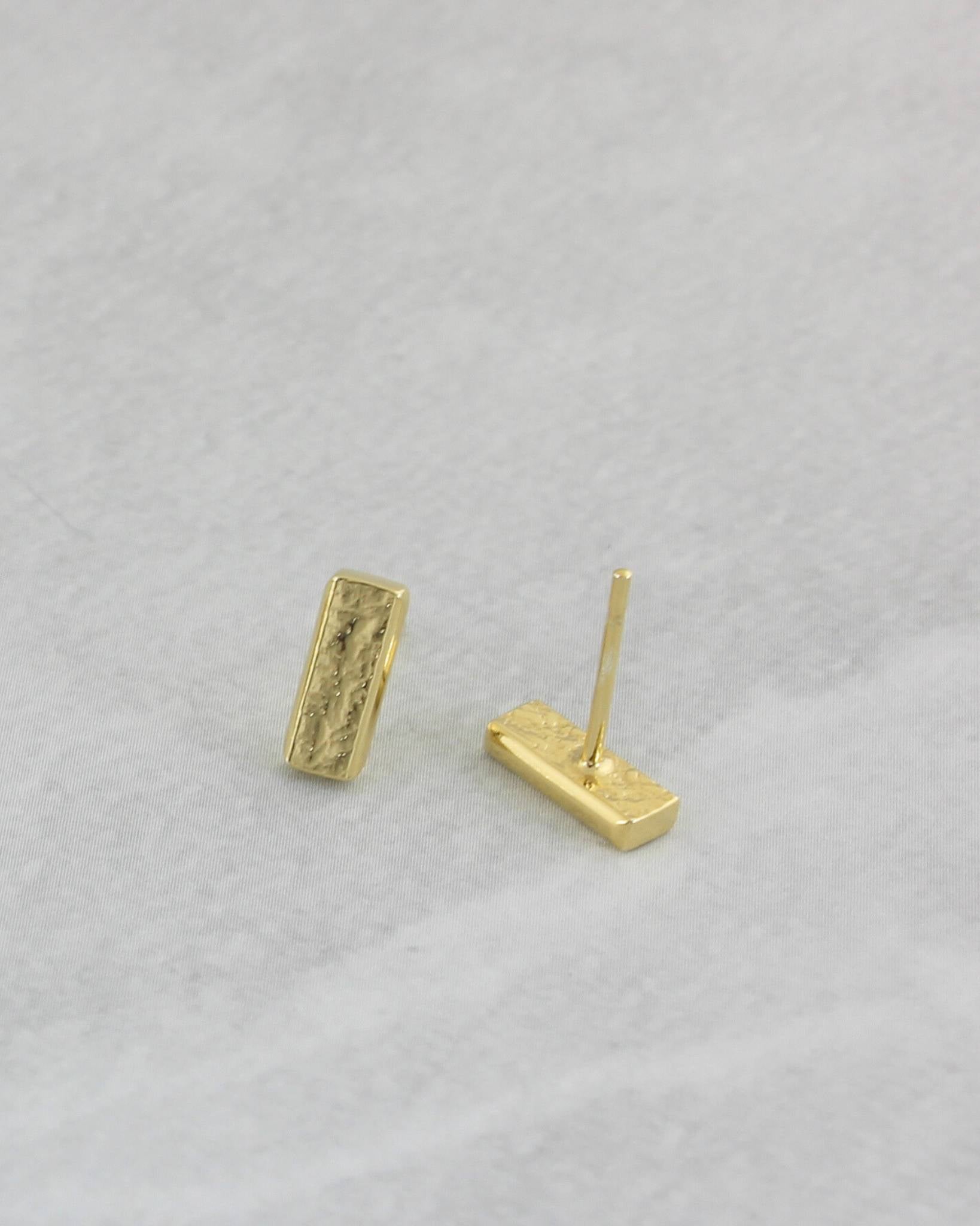 Dainty gold rectangle bar stud earrings with textured surface lying on light coloured marble paper