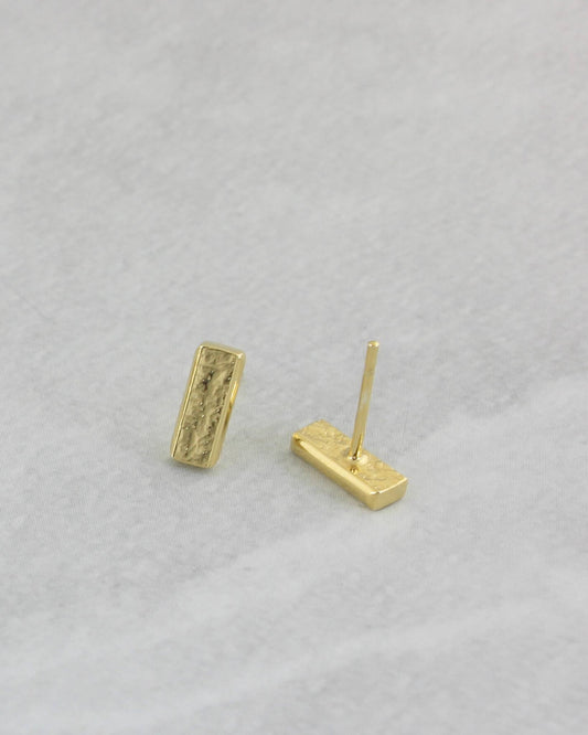 Dainty gold rectangle bar stud earrings with textured surface lying on light coloured marble paper