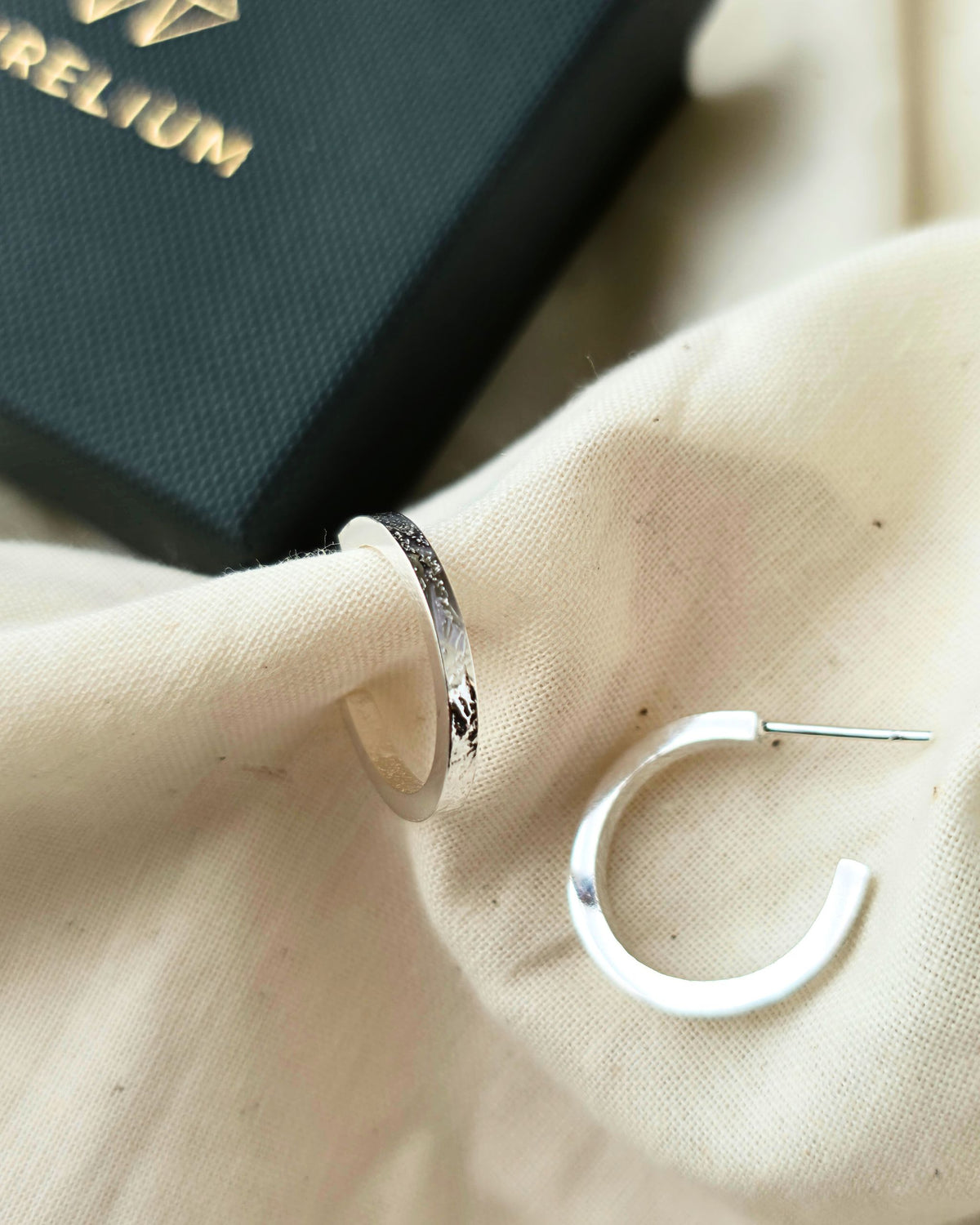 Textured silver hoop earrings on beige fabric with black Aurelium gift box in background