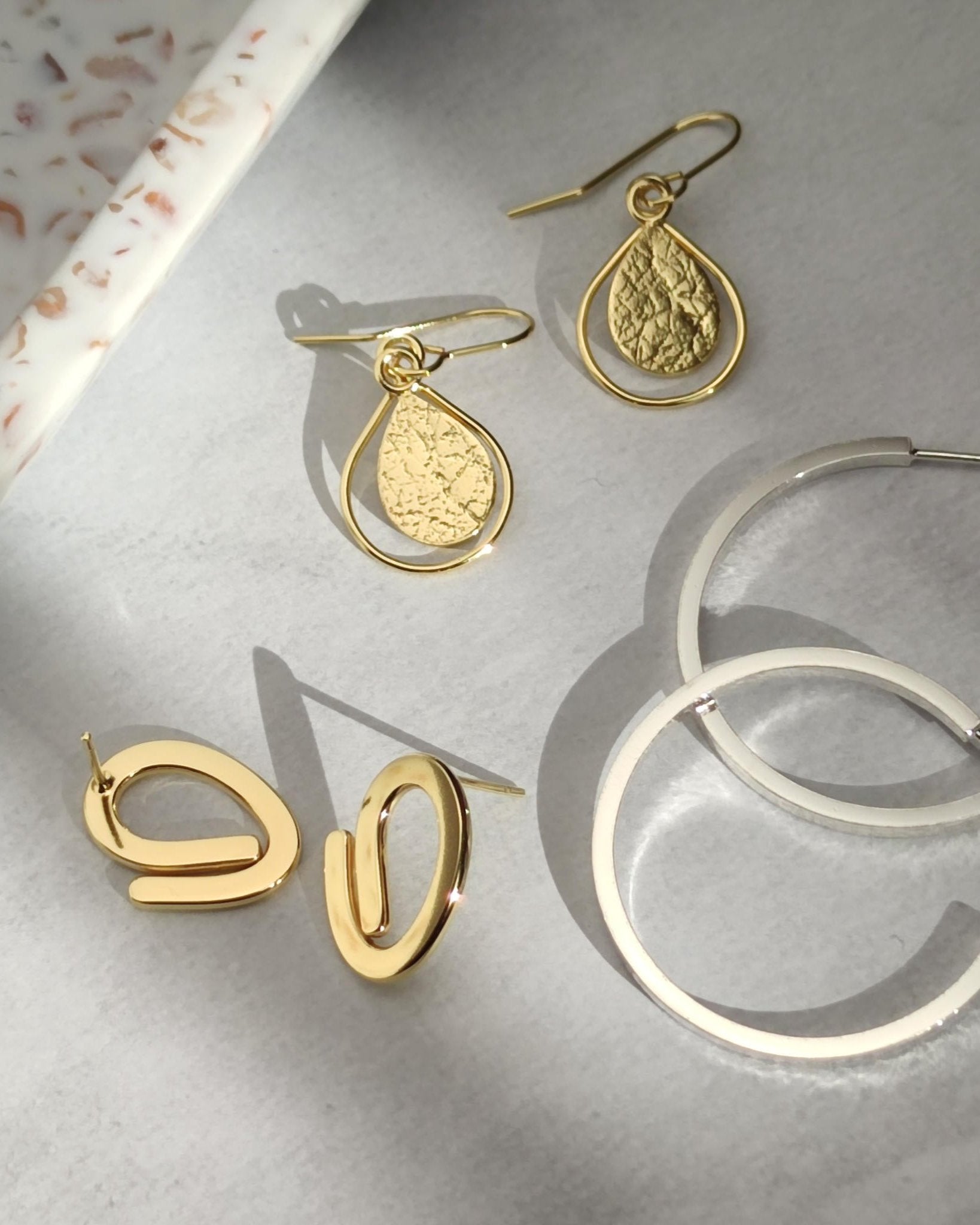 Three pairs of gold and silver handmade earrings lying in sunshine on grey surface beside speckled ceramic dish