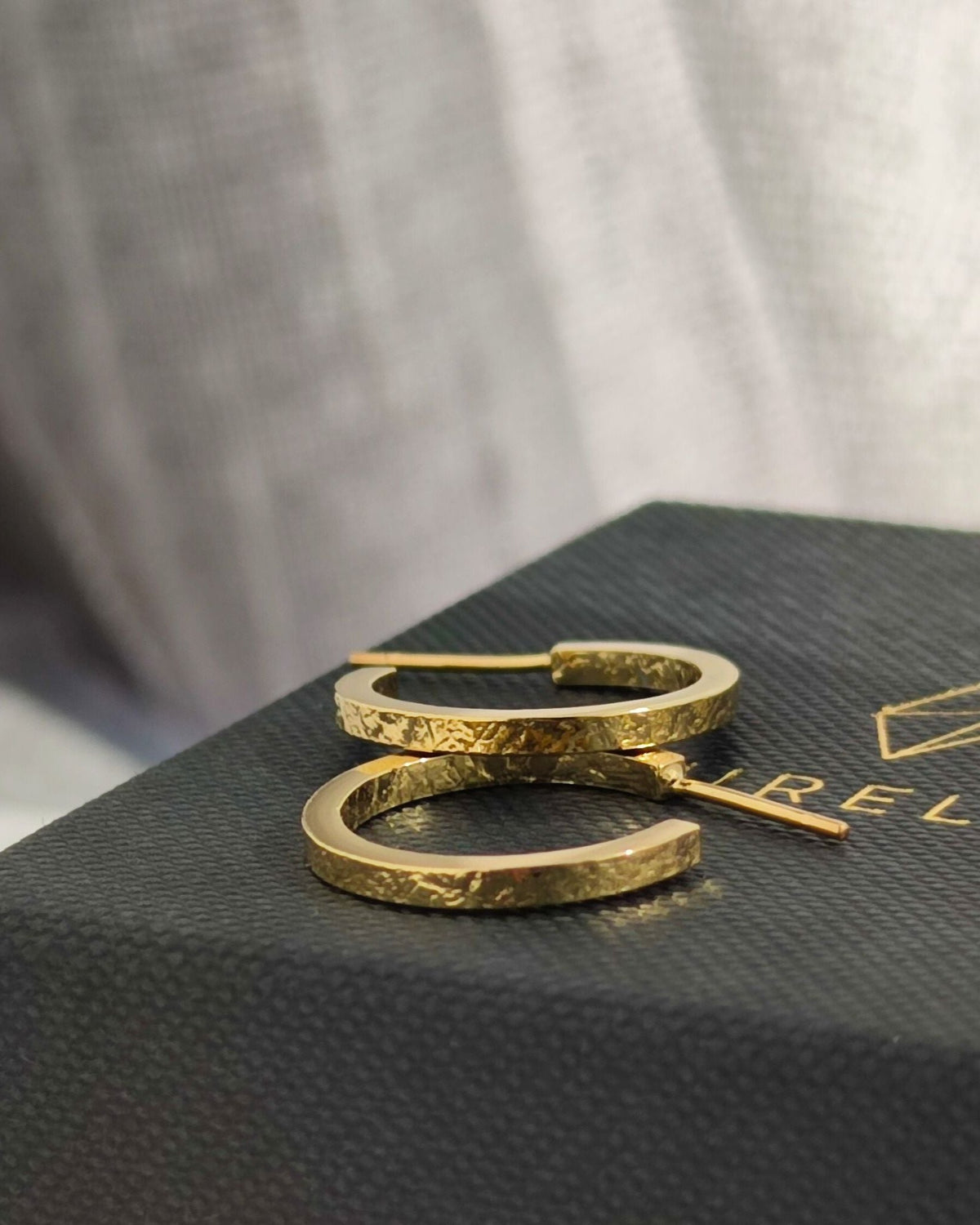 Gold hoop earrings with texture on front and back lying on closed black jewellery gift box with blurred background
