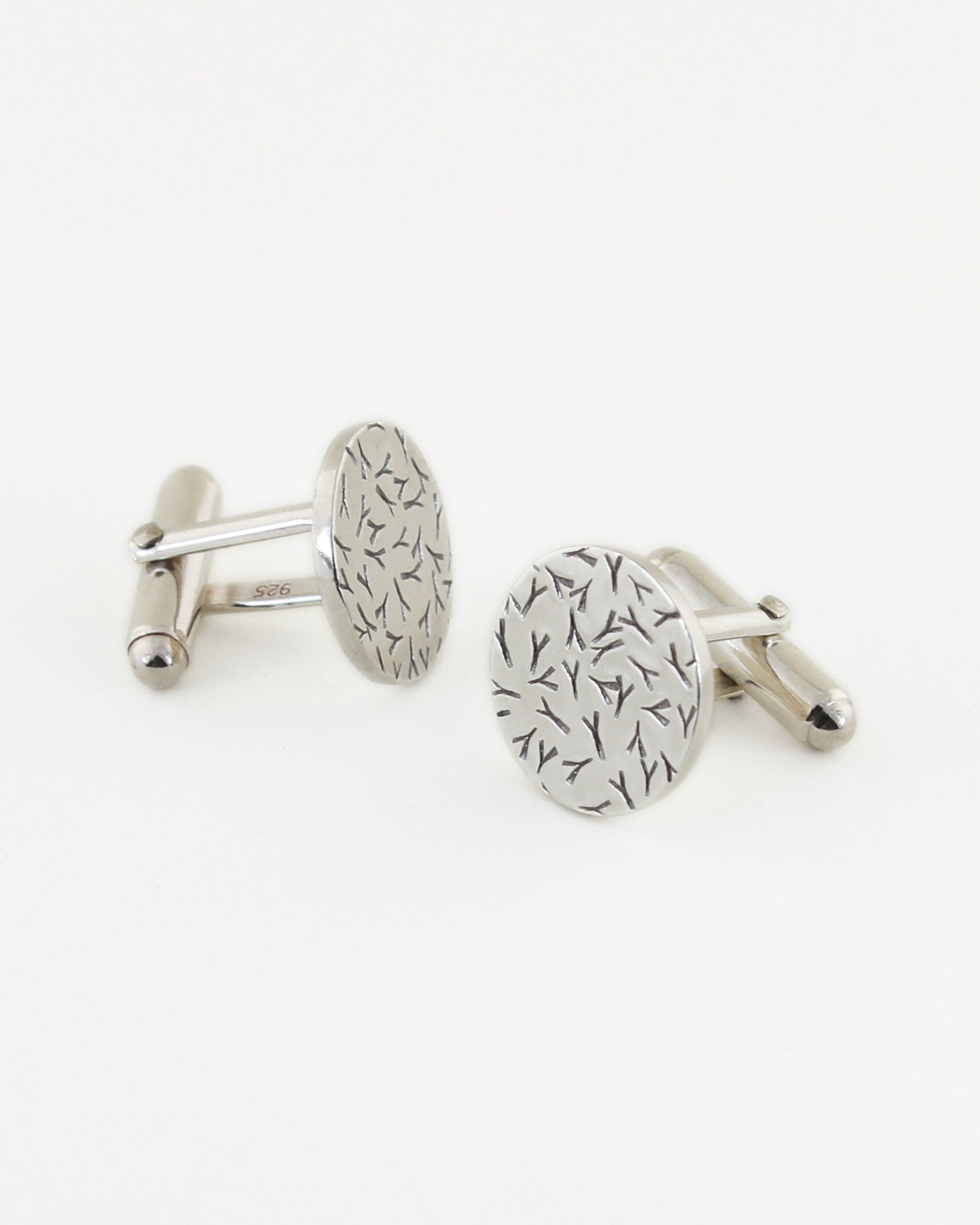 Front and side views of silver circle-shaped cufflinks with a contrasted silver and black textured surface, with white background