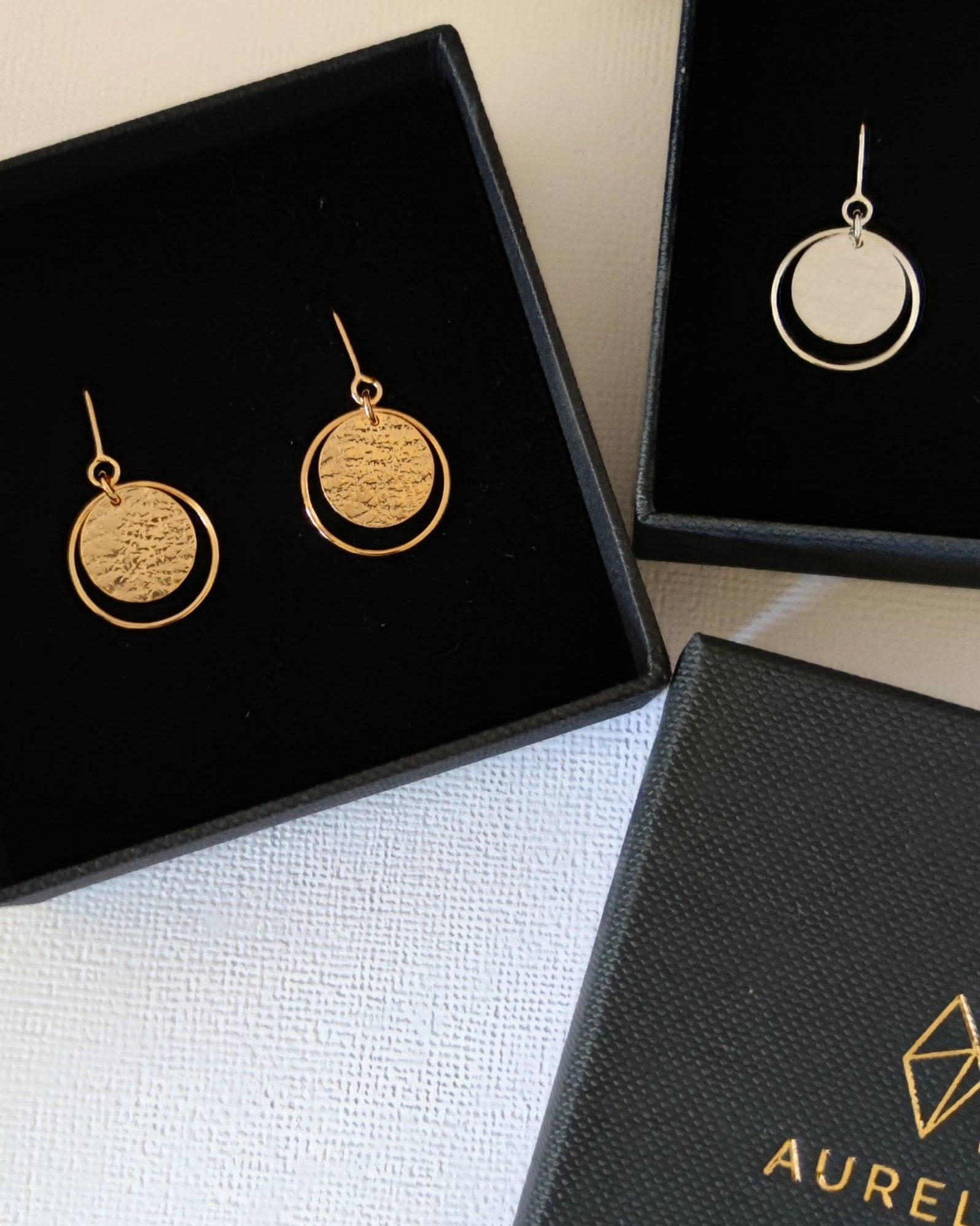 Delicate gold and silver earrings consisting of textured circles framed with thin wire loops and presented in open black jewellery gift boxes