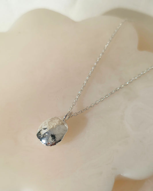 A silver chain necklace with a polished, oval-shaped silver pendant resting on a smooth, pale cream-coloured stone surface