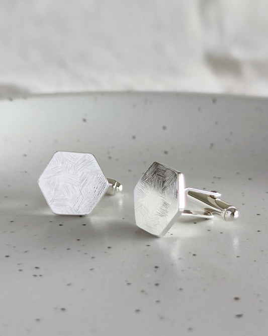 Handmade hexagon-shaped sterling silver cufflinks with a delicate textured surface lying on speckled cream ceramic dish