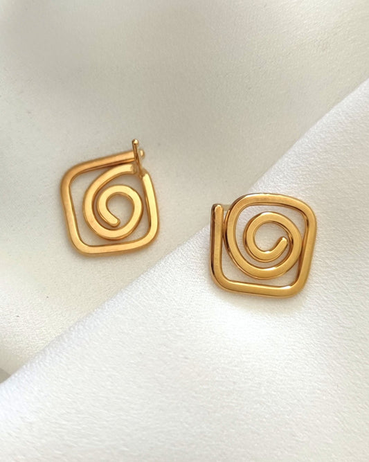 Small gold square-shaped stud earrings in swirl design lying on white fabric