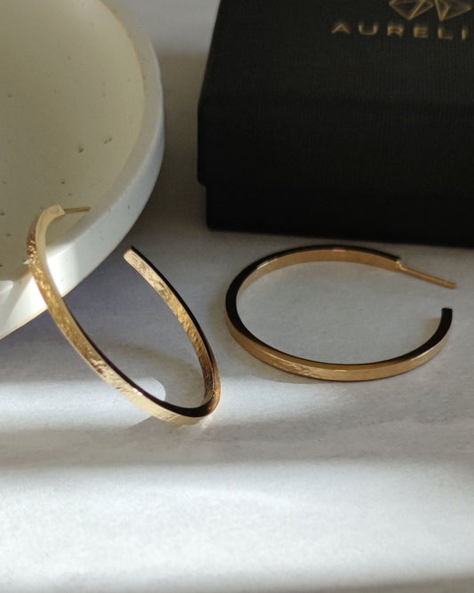 Large gold hoop earrings with texture printed on front and back lying on grey surface beside black gift box and ceramic plate