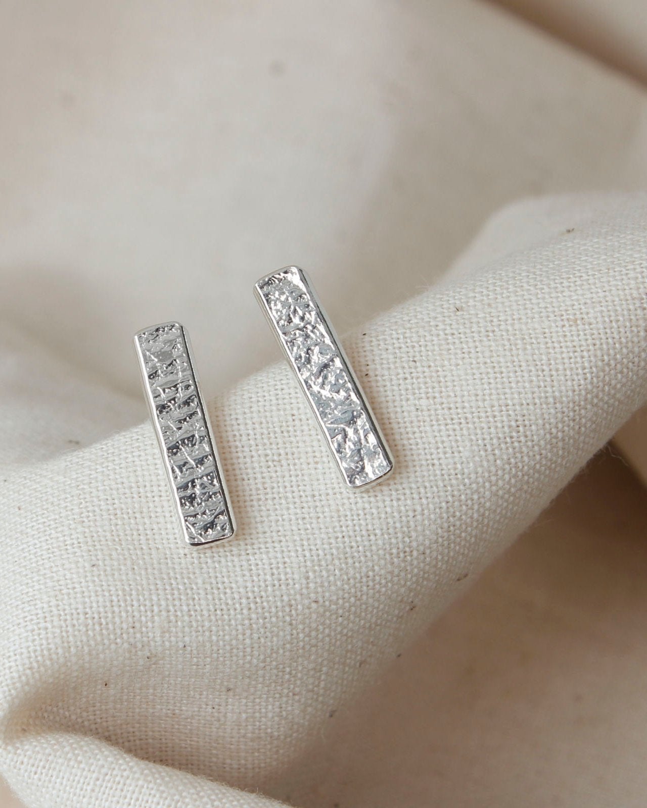 Minimalist long rectangle bar stud earrings with delicate textured surface lying on beige fabric