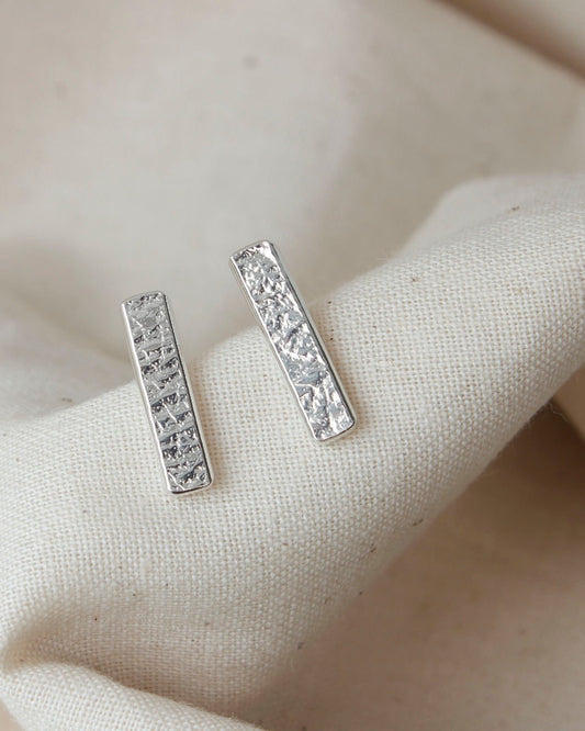 Minimalist long rectangle bar stud earrings with delicate textured surface lying on beige fabric