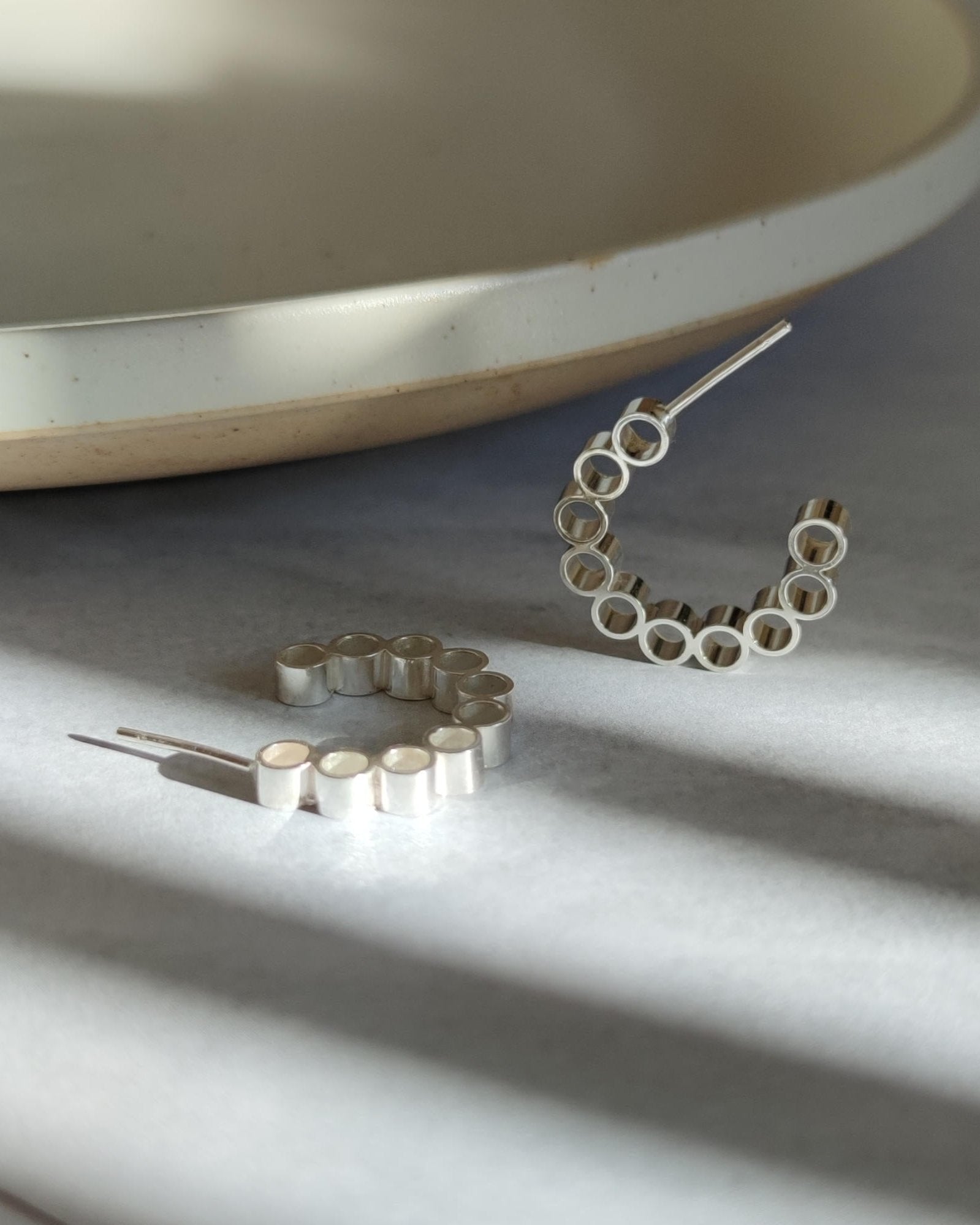 Handmade silver hoop earrings made from multiple pieces of tube lying in sunshine on grey background beside ceramic plate