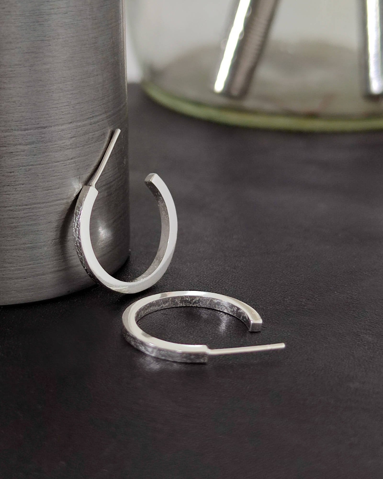 Small handmade silver hoop earrings with texture printed on front and back lying on dark surface with tools in background