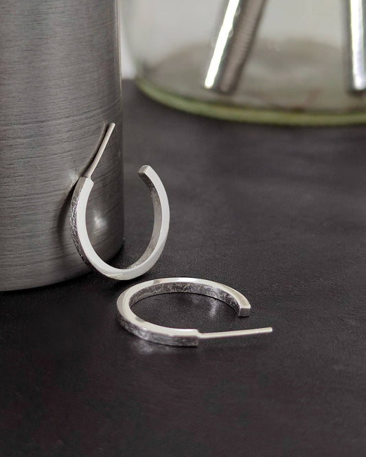 Small handmade silver hoop earrings with texture printed on front and back lying on dark surface with tools in background