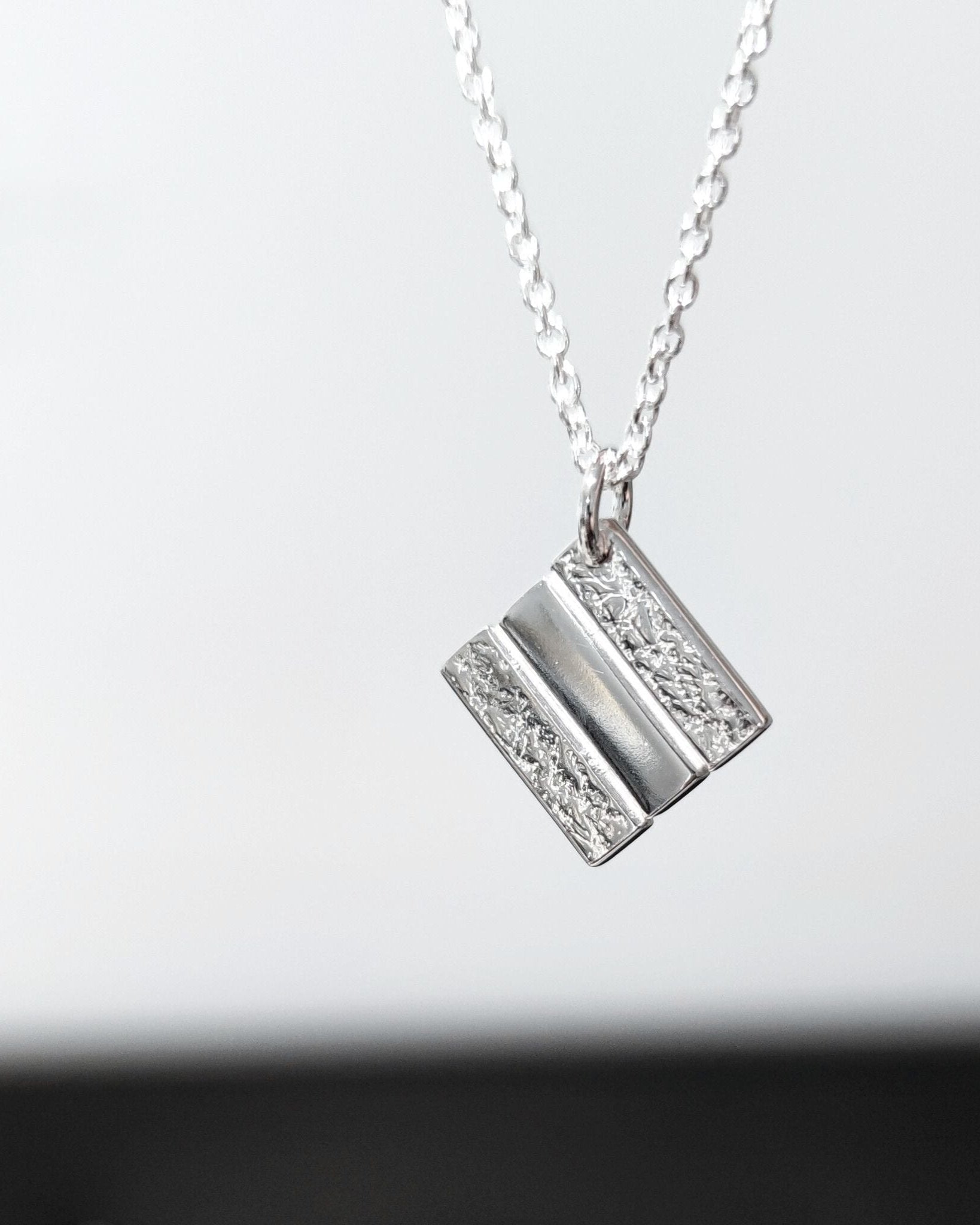 Handmade silver square pendant with partially textured surface hanging from delicate silver chain in front of blurred background