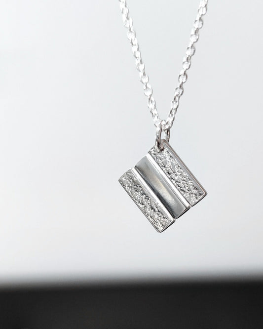 Handmade silver square pendant with partially textured surface hanging from delicate silver chain in front of blurred background