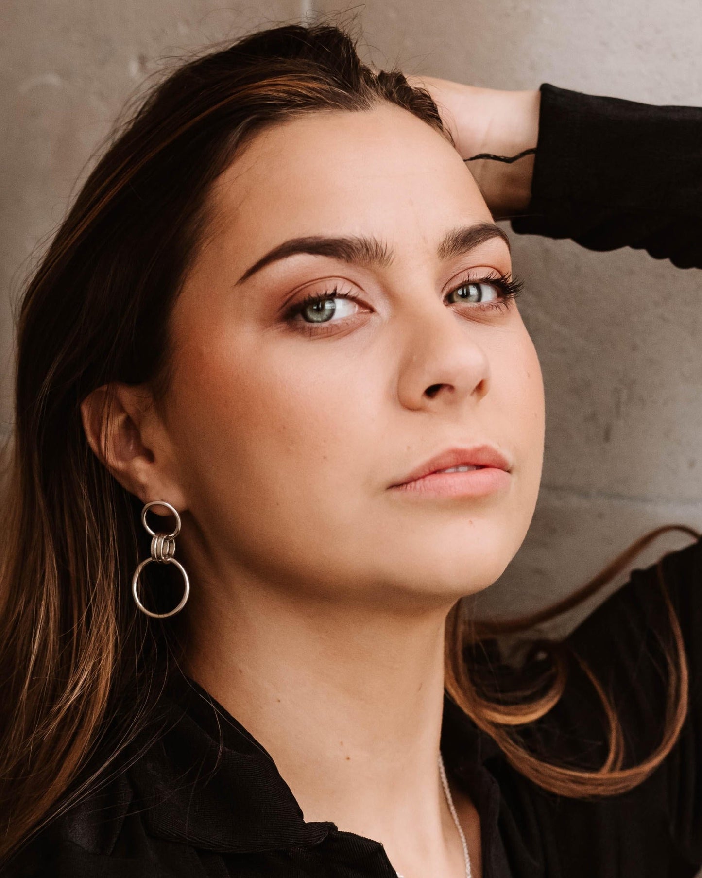 Person with long brown hair looking at camera and wearing dangly silver drop earrings made from small and large loops
