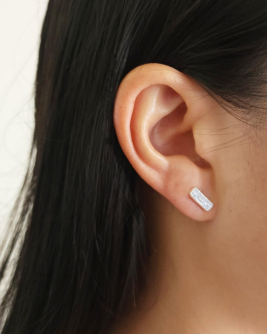 Person with black hair wearing small textured rectangle bar stud earrings in silver
