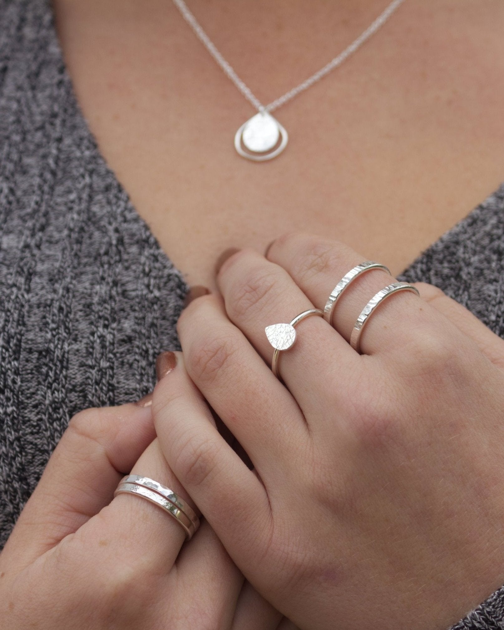 Person in grey jersey with hands cupped against chest, wearing handmade silver rings and teardrop-shaped pendant
