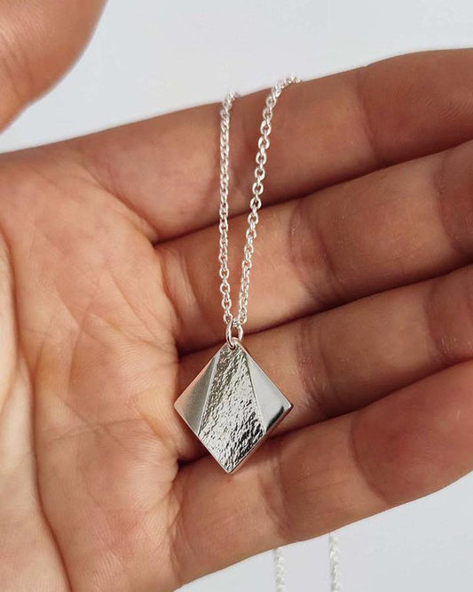 Close up of hand holding silver square pendant with partially textured surface hanging on delicate silver chain
