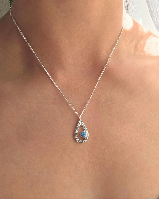 Silver teardrop necklace with a textured surface set with a blue topaz gemstone on a person's neck