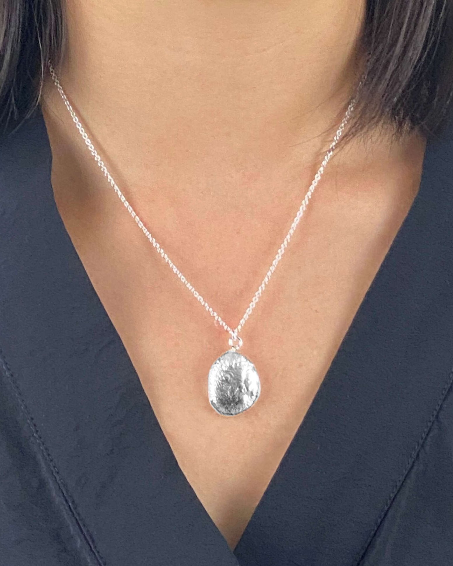 Front view of person wearing oval-shaped molten silver pendant with reflective rippled texture hanging on fine silver chain