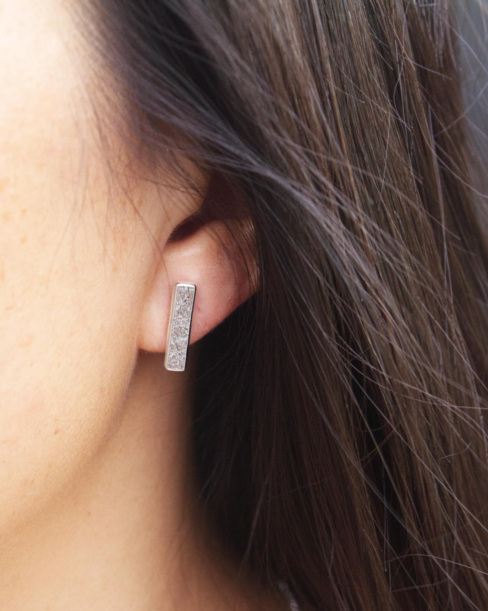 Person with dark brown hair wearing silver rectangle bar stud earring with textured surface