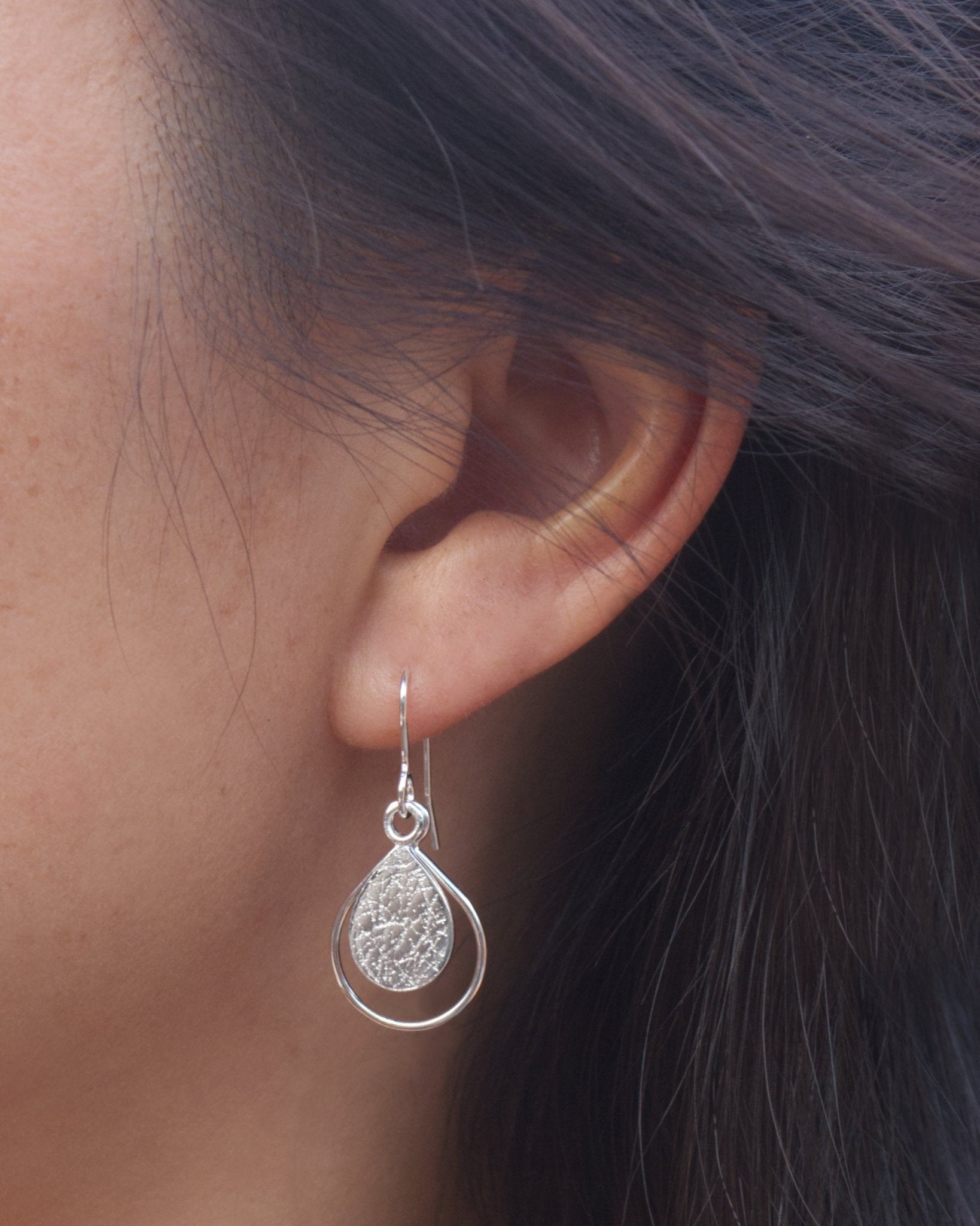 Close up of person with black hair wearing handmade silver earrings with textured teardrop, framed by thin wire teardrop