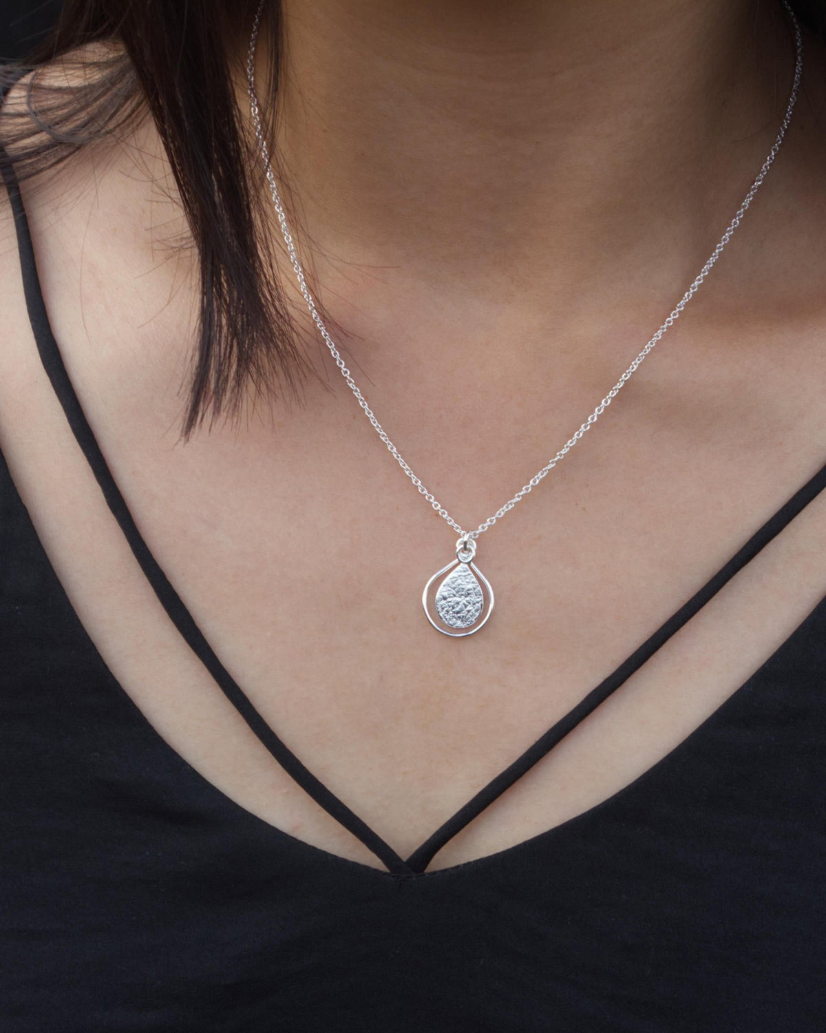 Front view of person in black top wearing textured silver teardrop pendant framed by thin silver wire teardrop, hanging from delicate silver chain