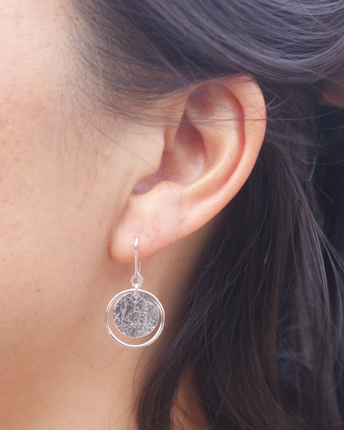 Close up of person wearing handmade silver earrings consisting of a textured circle framed by a thin silver wire loop