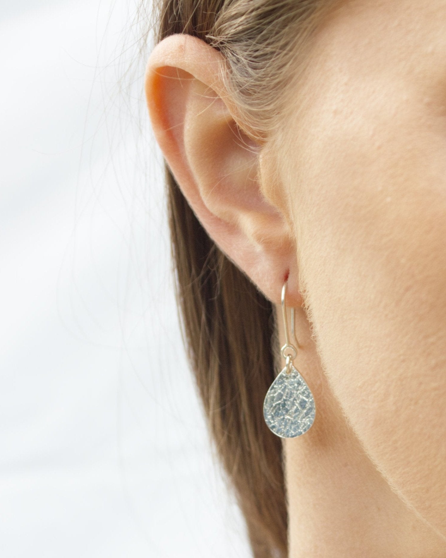 Close up of person wearing teardrop-shaped silver earrings with delicate texture against white backdrop