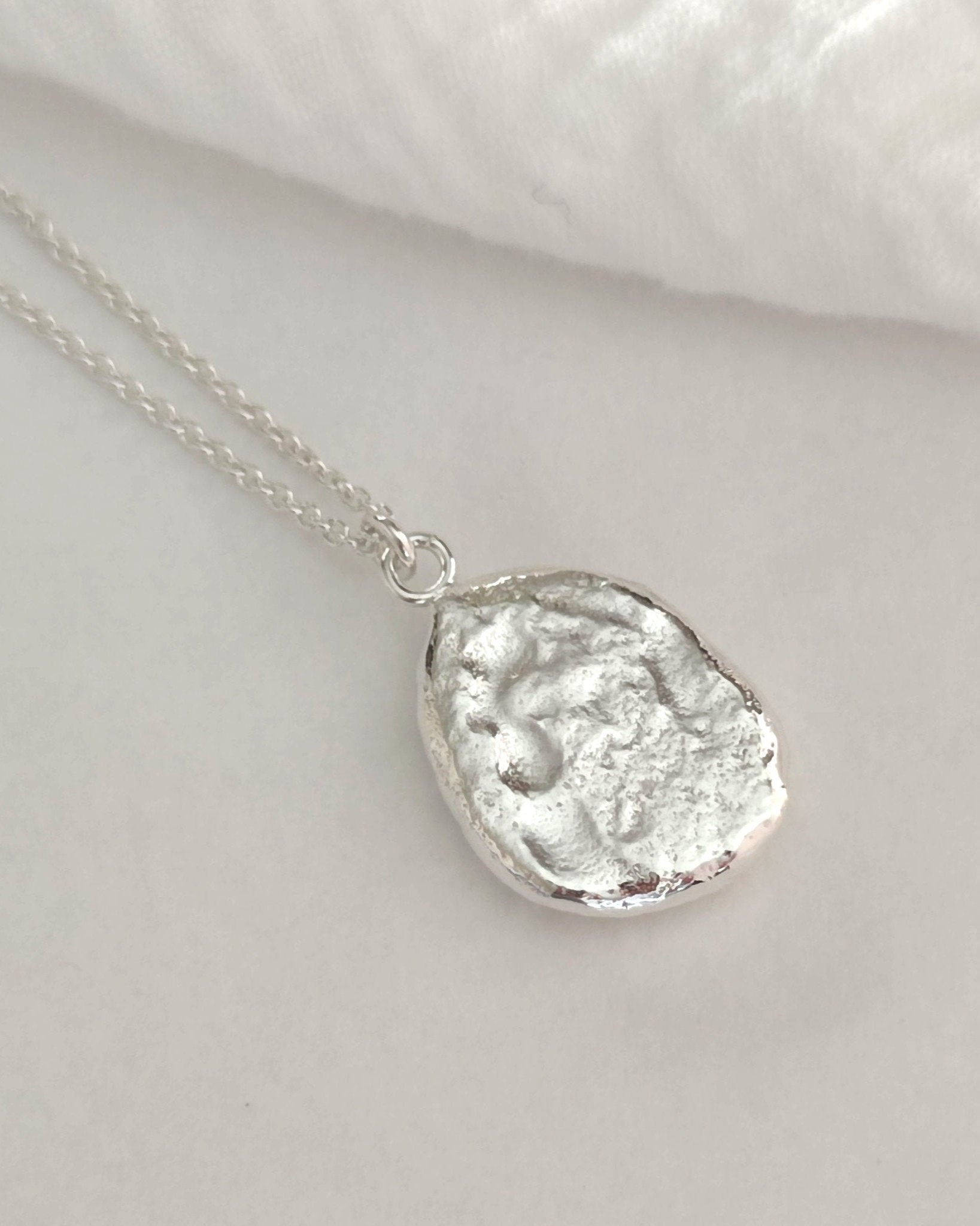Back view of chunky molten silver pendant with smooth textured surface on silver cable chain and lying on white fabric