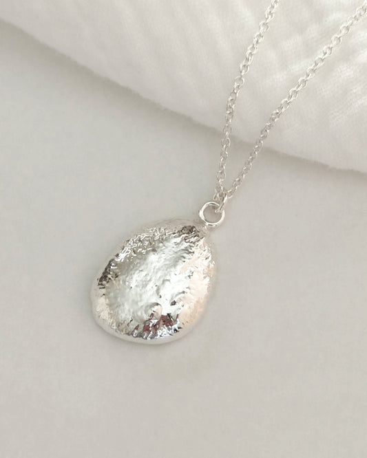 Chunky molten silver pendant in oval shape on silver cable chain and lying on white fabric