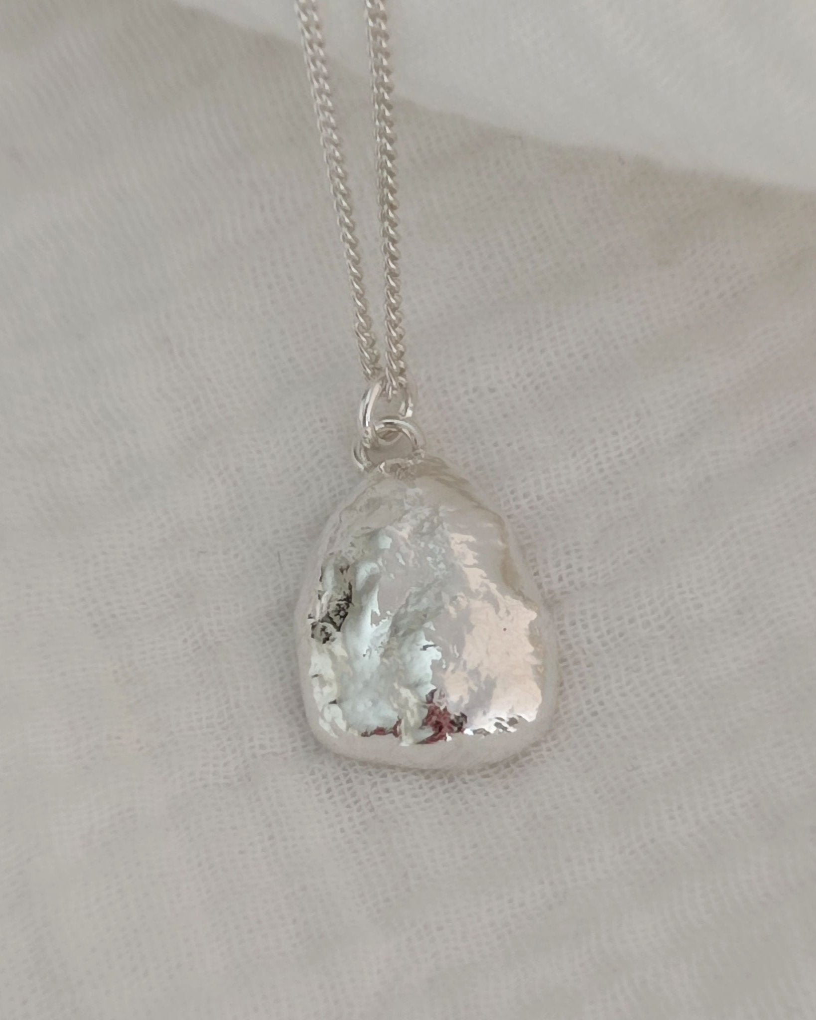 Molten silver pendant with organic texture attached to fine silver chain and lying on soft white fabric
