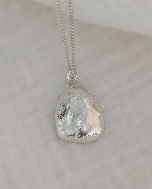 Molten silver pendant with organic texture attached to fine silver chain and lying on soft white fabric