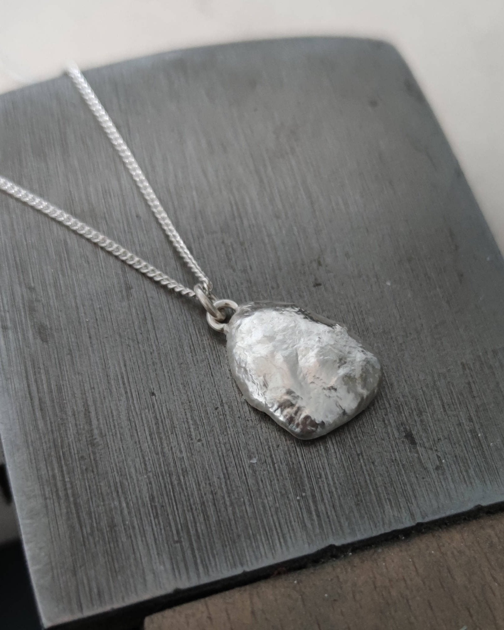 Molten silver pendant with reflective rippled surface texture attached to fine silver chain and lying on jewellery workbench