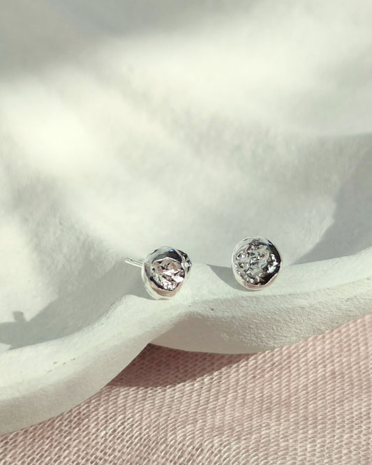 Small circle-shaped molten silver studs with organic texture lying on white ceramic dish on top of pink fabric