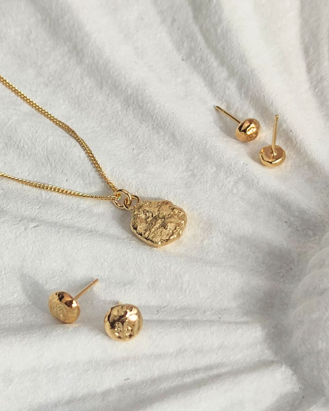 Two pairs of gold circle-shaped stud earrings and one small gold pebble-shaped pendant lying on white ceramic dish