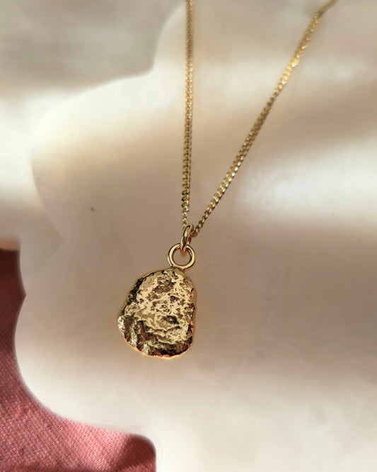 Gold necklace wit h a textured pebble pendant lying in diffused sunlight on a cream background