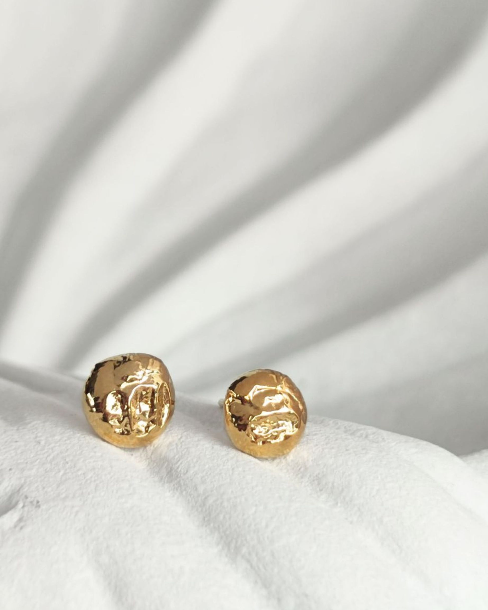 Handmade nugget-shaped molten gold stud earrings with an organic rippled texture lying on white ceramic shell