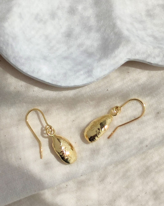 Molten gold drop earrings with organic aesthetic lying on fabric in sunshine beside white ceramic dish