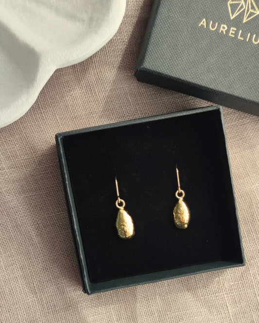 Elegant gold teardrop-shaped earrings in black jewellery gift box lying on beige fabric with box lid with the word "Aurelium" in gold lettering nearby