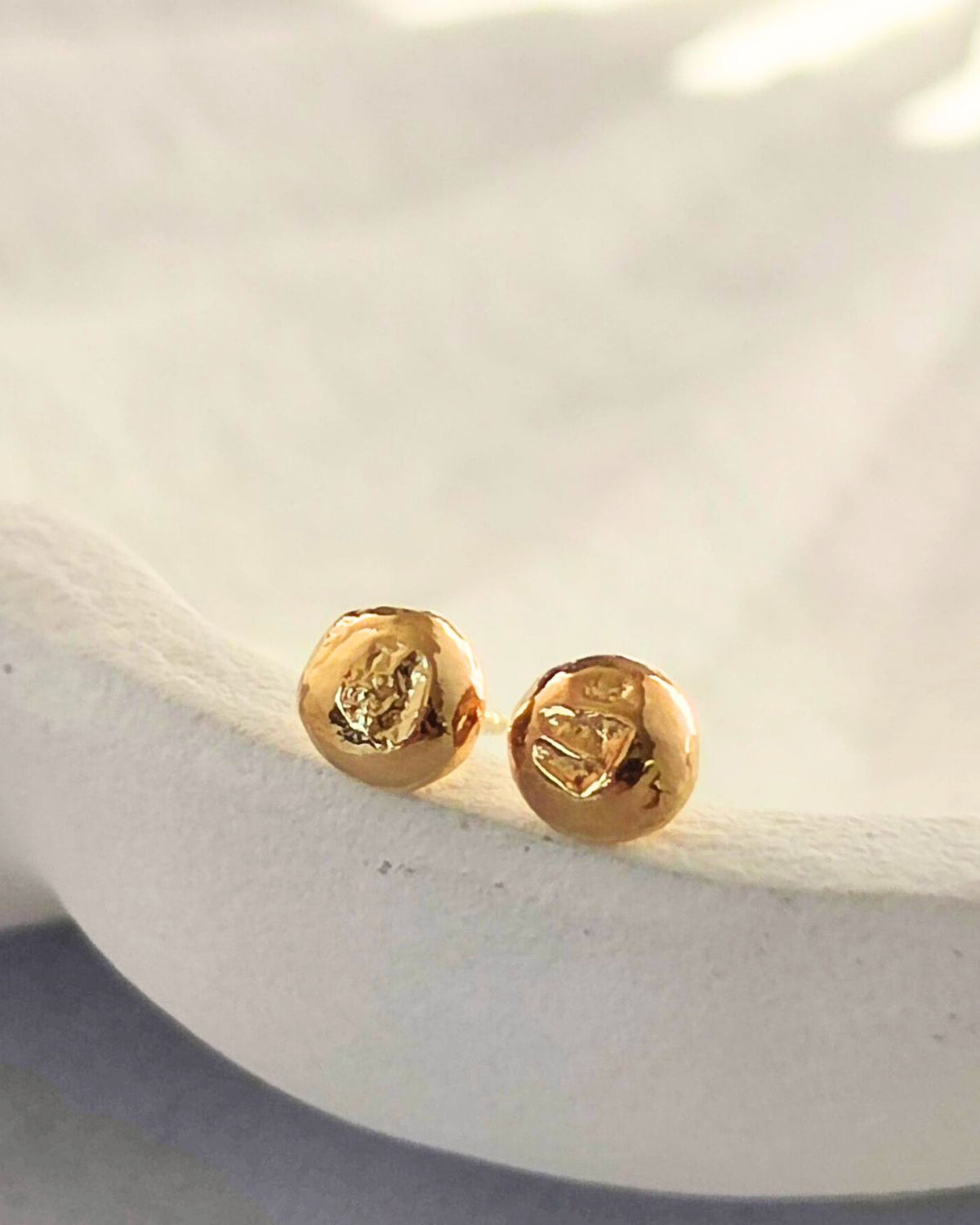 small molten gold circle shaped stud earrings with textured surface lying on cream dish