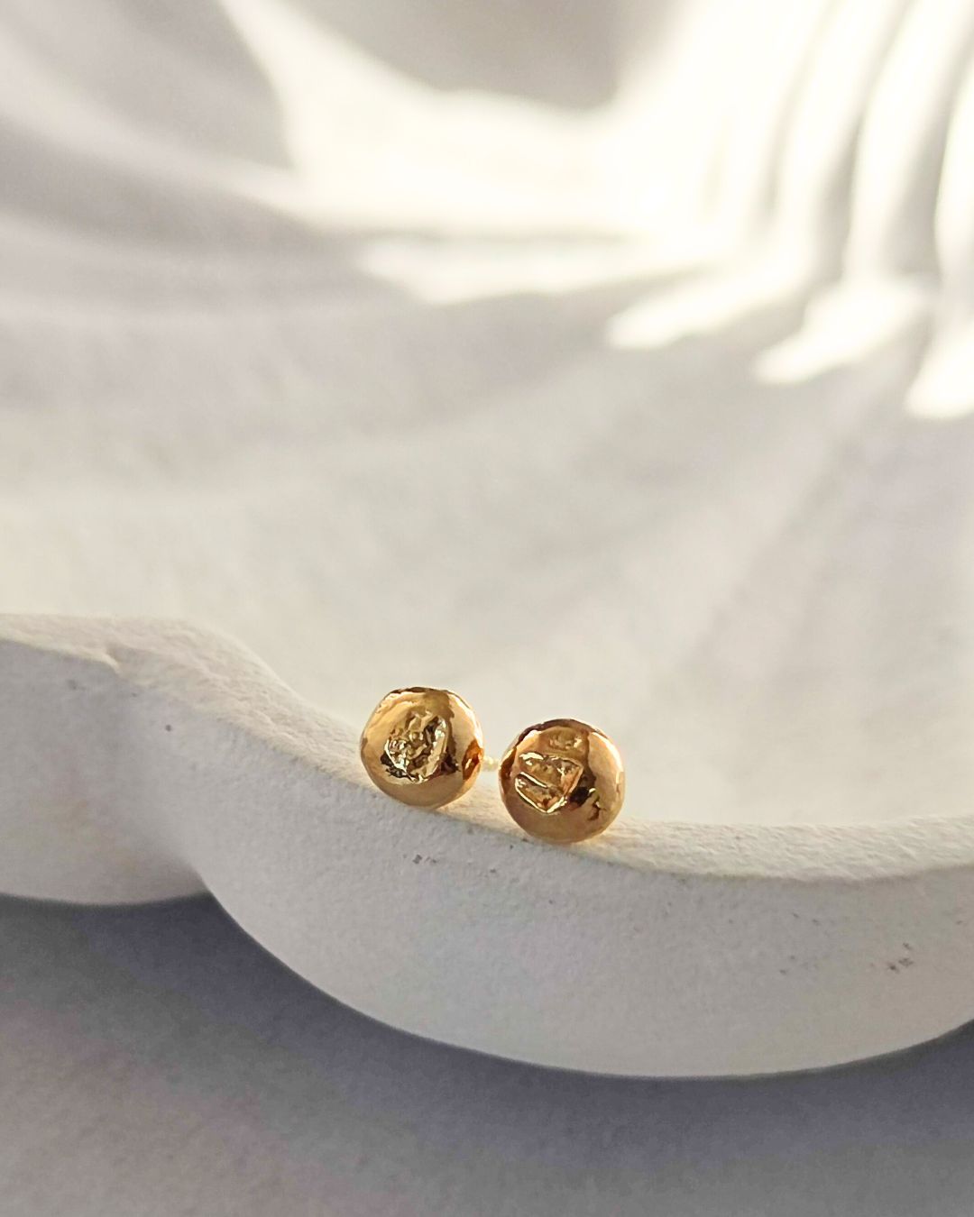 Organic molten gold stud earrings with unique surface texture lying on ceramic dish