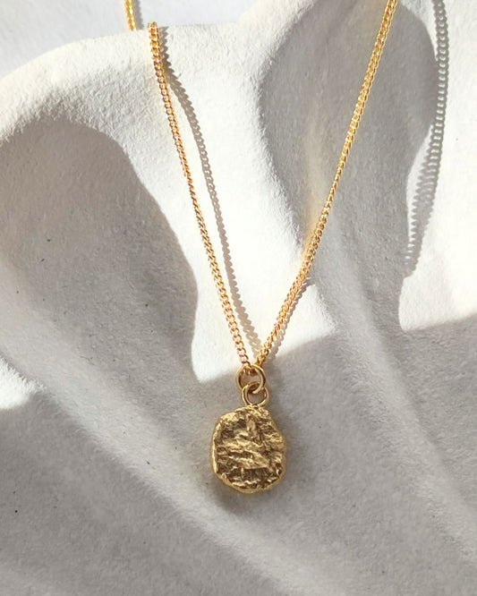 Handmade molten gold pendant with a pebble-shaped formation lying in shadow on sunlit white ceramic dish